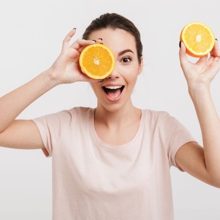 A woman in a light pink shirt holds two orange halves, one covering her eye, and smiles against a plain white background.