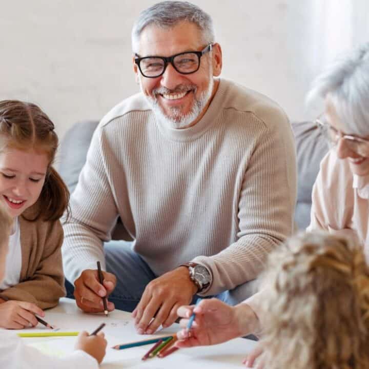 Two adults and three children sit around a table drawing with colored pencils, smiling and engaging in arts and crafts together in a well-lit room.