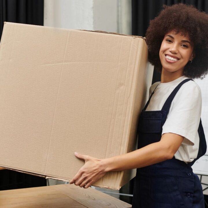 A person in overalls smiles while holding a large cardboard box indoors, with other boxes and a flipchart in the background.