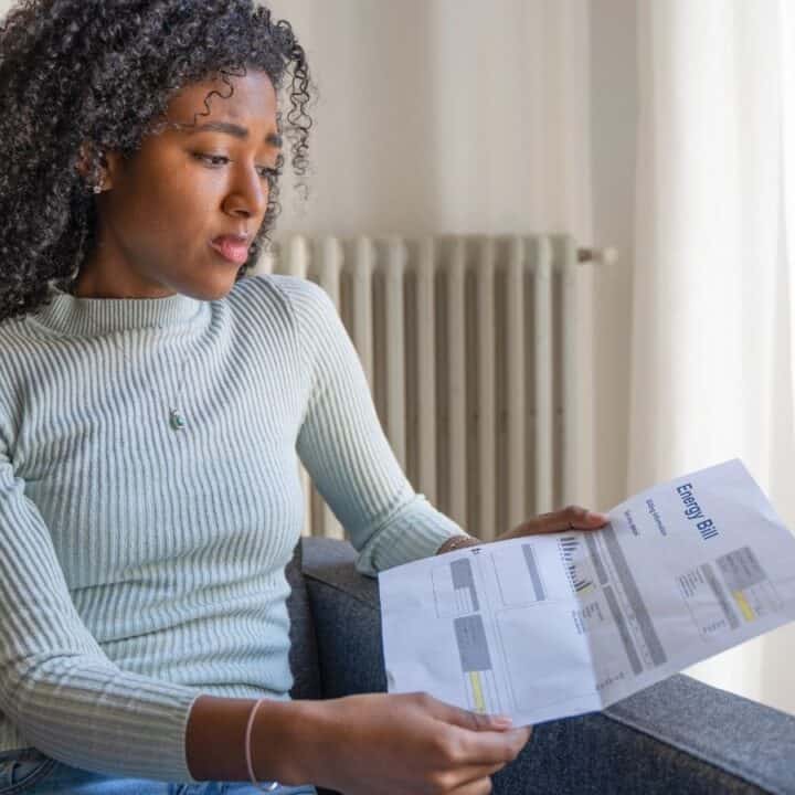 A woman sits on a couch, looking at an energy bill she is holding in her hands.