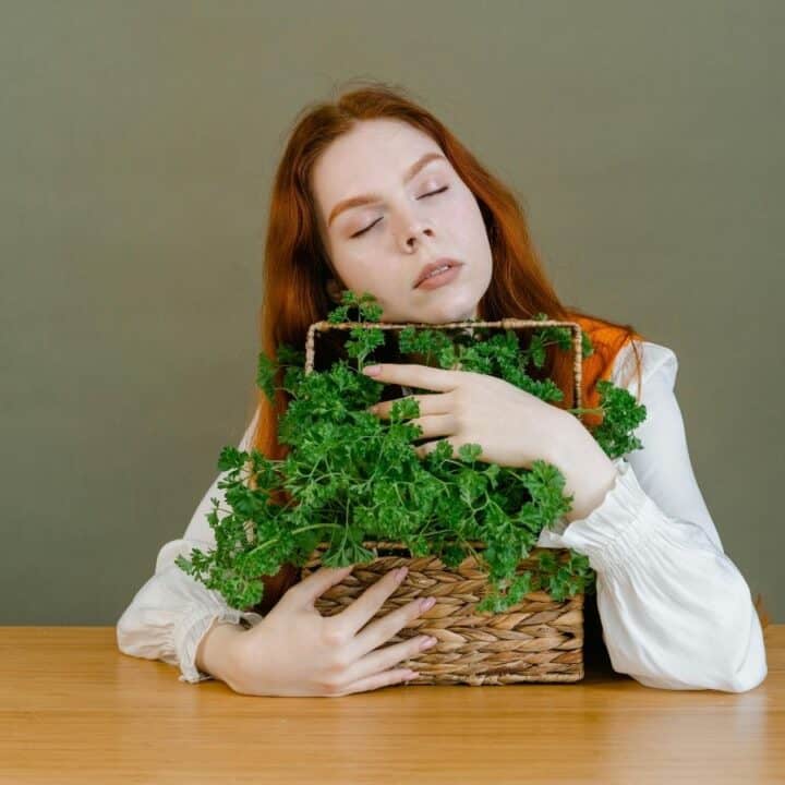A person with long red hair sits at a table, eyes closed, holding a woven basket filled with leafy green herbs, against a plain background.