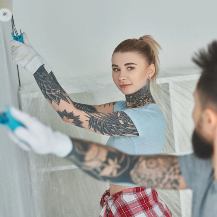 Two people with tattoos and gloves are painting a wall with white paint rollers. Plastic sheets cover furniture in the background.