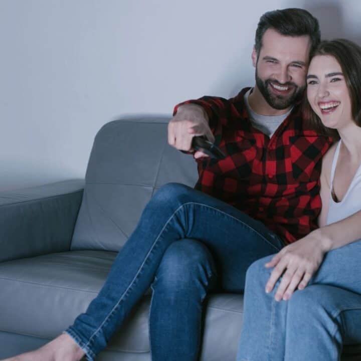 A man and woman sit together on a gray couch, smiling and watching TV, with the man holding a remote control.