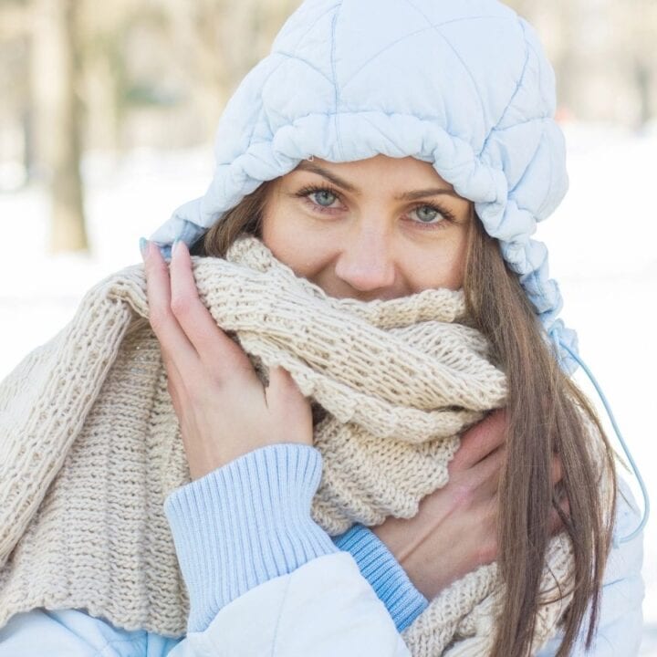 A woman outdoors in winter wears a blue hooded jacket and beige knit scarf, holding the scarf close to her face with both hands. Snow covers the background.