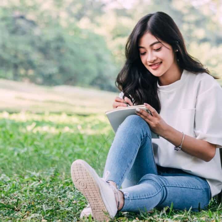 A young woman sits on grass in a park, writing in a notebook with a backpack beside her.