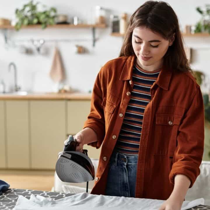 A woman stands in a kitchen, ironing a white shirt on an ironing board. She wears a rust-colored jacket over a striped shirt.
