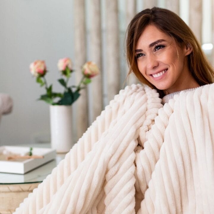 A woman sits on a couch wrapped in a cream-colored blanket, smiling. A round table with a vase of flowers and a tray is in the background.