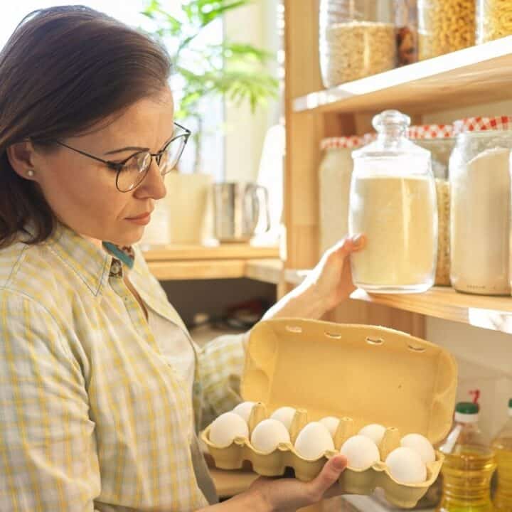 A woman holding a carton of eggs stands in front of shelves stocked with jars of dry goods in a well-lit pantry.