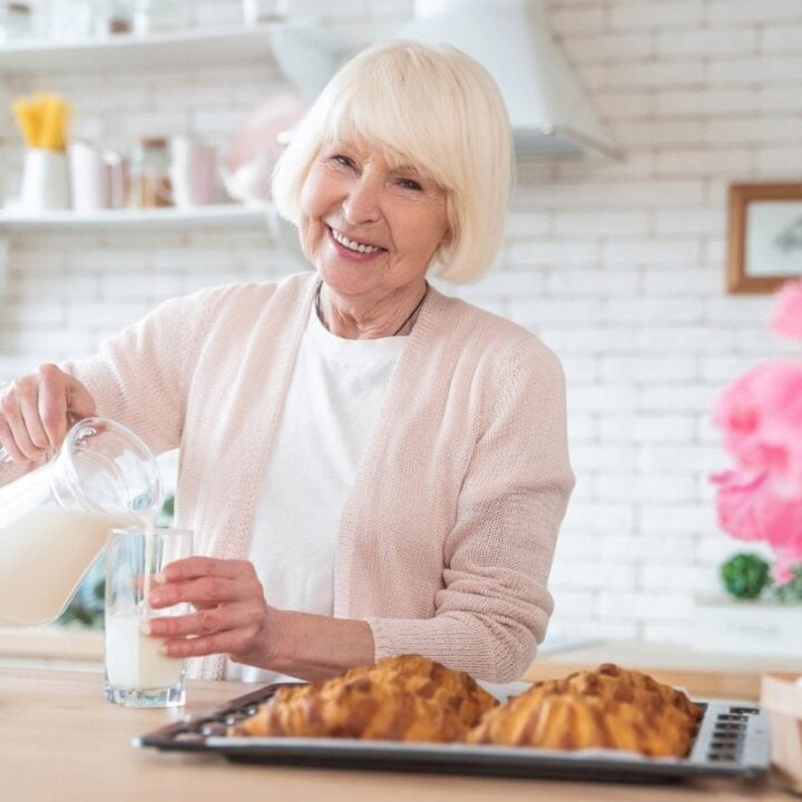 An older woman in a bright kitchen pours milk into a glass, surrounded by cookies, pastries, and pink flowers on the counter.