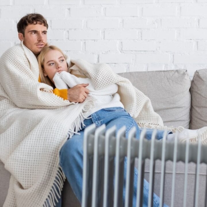 A man and woman sit on a couch wrapped in a blanket, looking at a heater in front of them.
