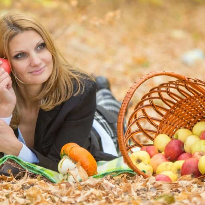 A woman lies on a blanket in fallen leaves, holding an apple. A tipped-over basket with apples is beside her.