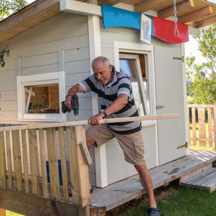 An older man uses a power drill to build a wooden railing on the porch of a small, light-colored playhouse in a grassy yard.
