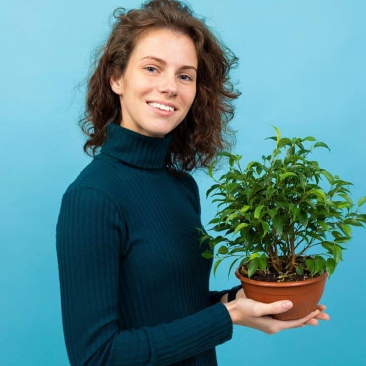 A woman in a dark turtleneck holds a potted green plant and smiles at the camera against a blue background.