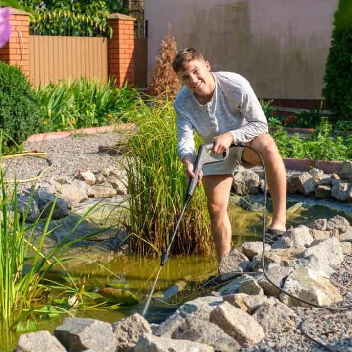 A young man uses a hose to clean or fill a small backyard pond surrounded by rocks and plants on a sunny day.