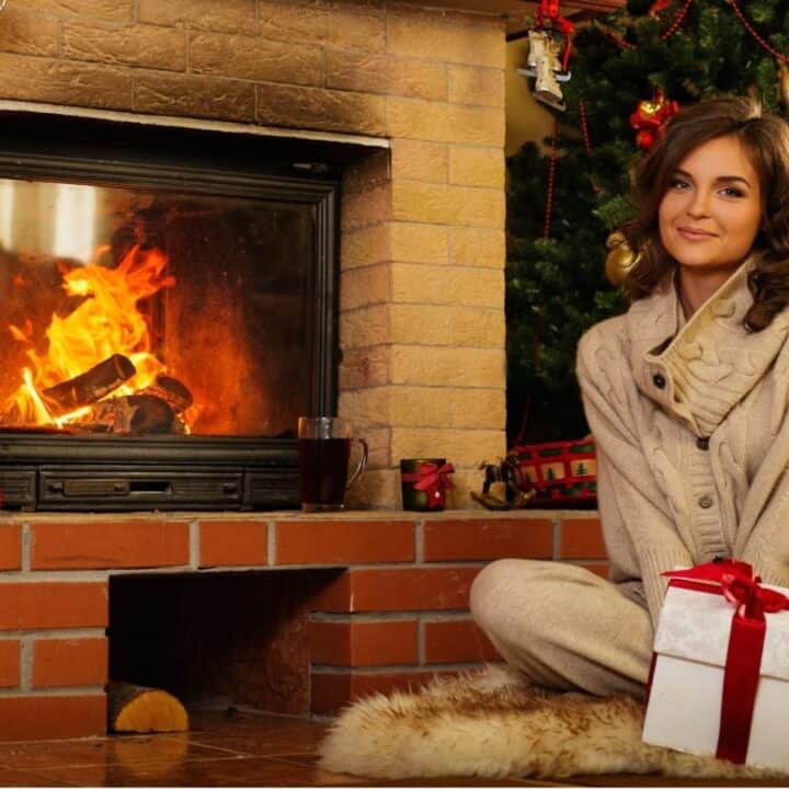 Woman in beige sweater sits on rug holding a wrapped gift in front of a fireplace and decorated Christmas tree.