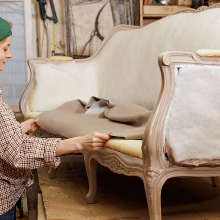 A person in a plaid shirt and green cap reupholsters a vintage sofa, pulling fabric over the seat in a workshop setting.