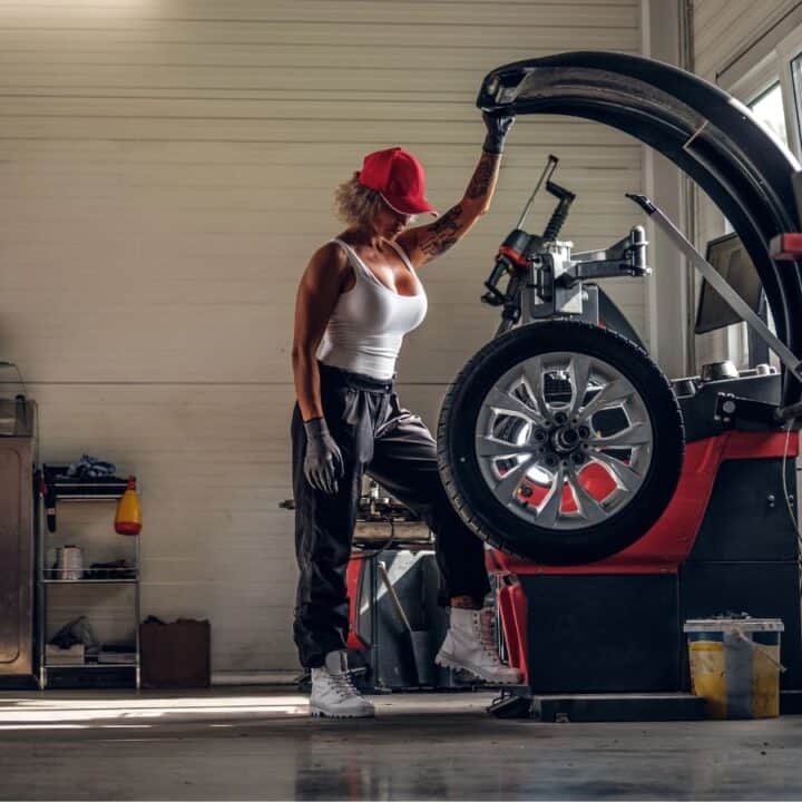 A woman in work clothes and a red cap operates a tire balancing machine in a well-lit auto repair workshop.
