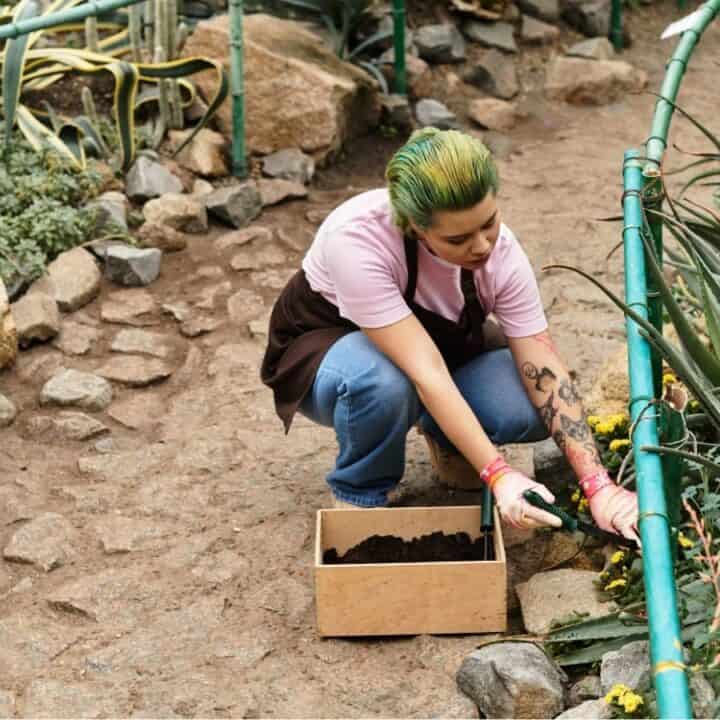 A person with green hair kneels on a dirt path in a garden, tending to plants with a small shovel and a box of soil nearby.