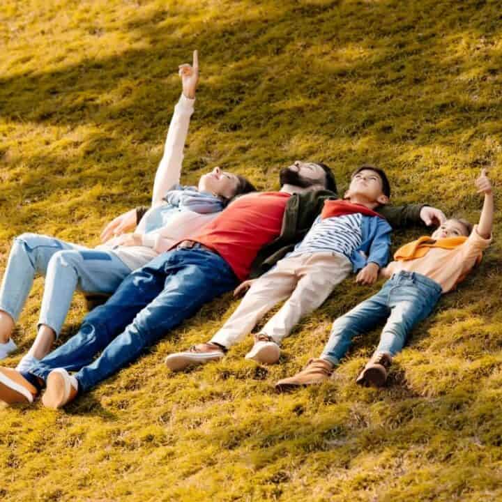 Four children lie on their backs on a grassy hill, looking up at the sky, with two of them pointing upwards.