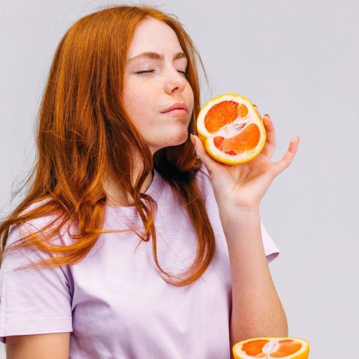 A young woman with long red hair holds half a grapefruit near her face with her eyes closed, wearing a light purple t-shirt against a plain background.