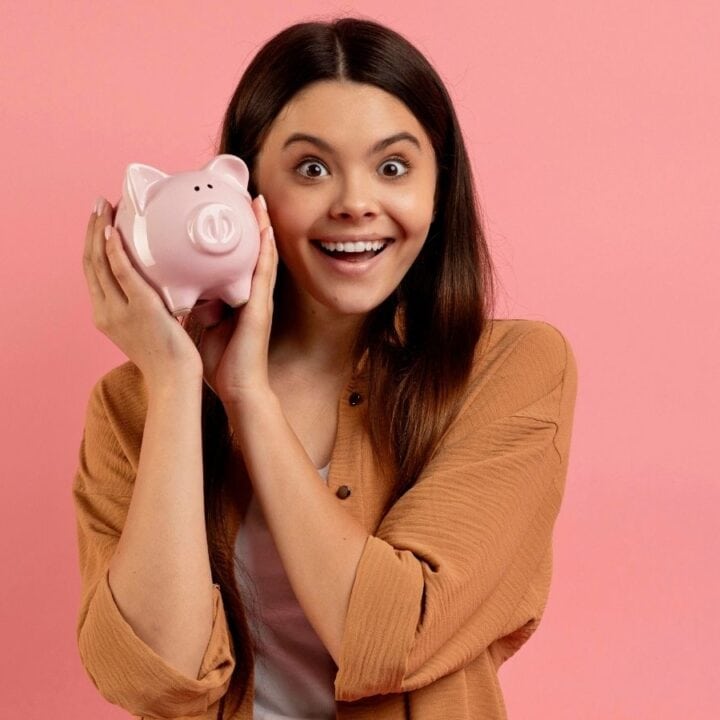 A woman in a brown jacket holds a pink piggy bank near her face and smiles, standing in front of a plain pink background.