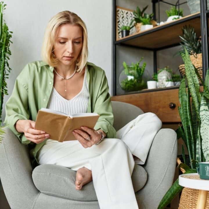 A woman sits in a gray chair reading a book, surrounded by various potted plants in a bright, modern indoor setting.