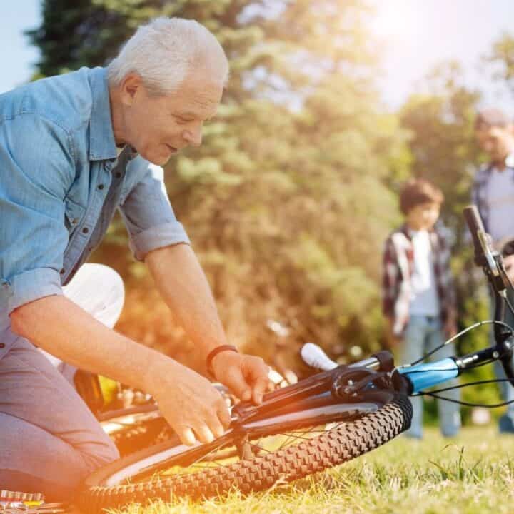 An older man kneels on grass repairing a bicycle, while two people, an adult and a child, stand in the background on a sunny day.