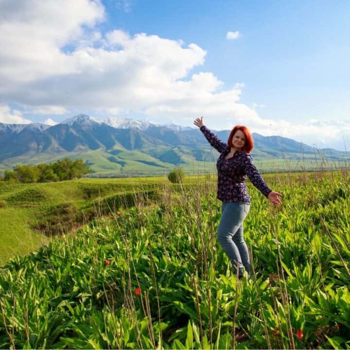 A woman stands with arms outstretched in a green field with mountains and a partly cloudy sky in the background.