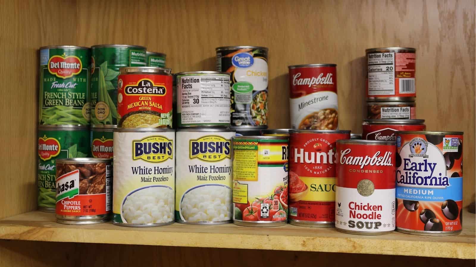 A wooden shelf stocked with various canned foods, including vegetables, soups, beans, corn, tomato sauce, olives, and peppers from multiple brands.