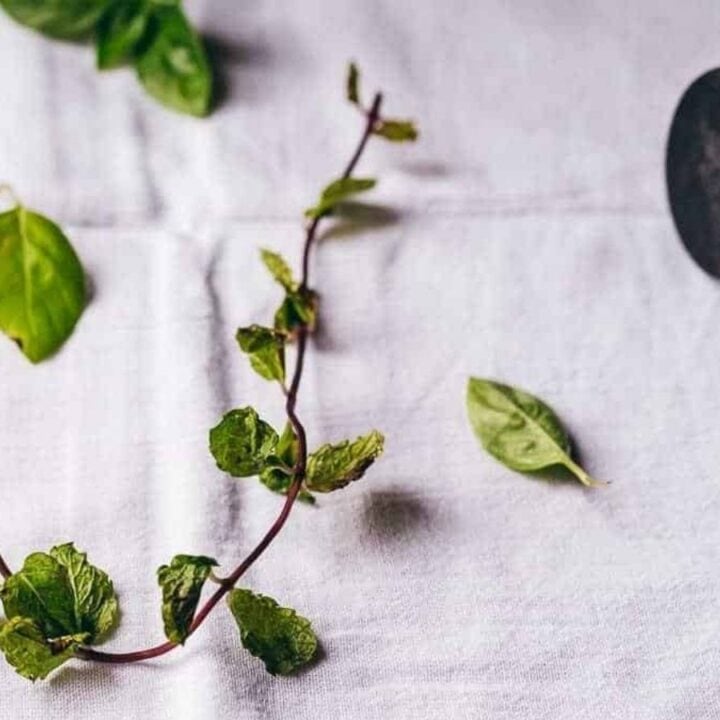 A sprig of mint and scattered basil leaves on a white cloth beside a black mallet with a wooden handle.