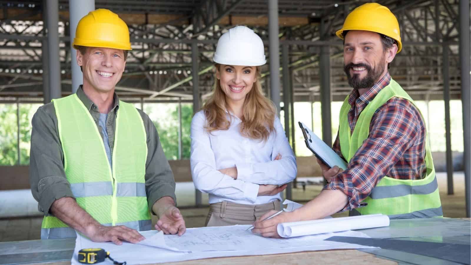Three construction professionals wearing safety helmets and vests stand at a table with blueprints and a clipboard inside a building under construction.