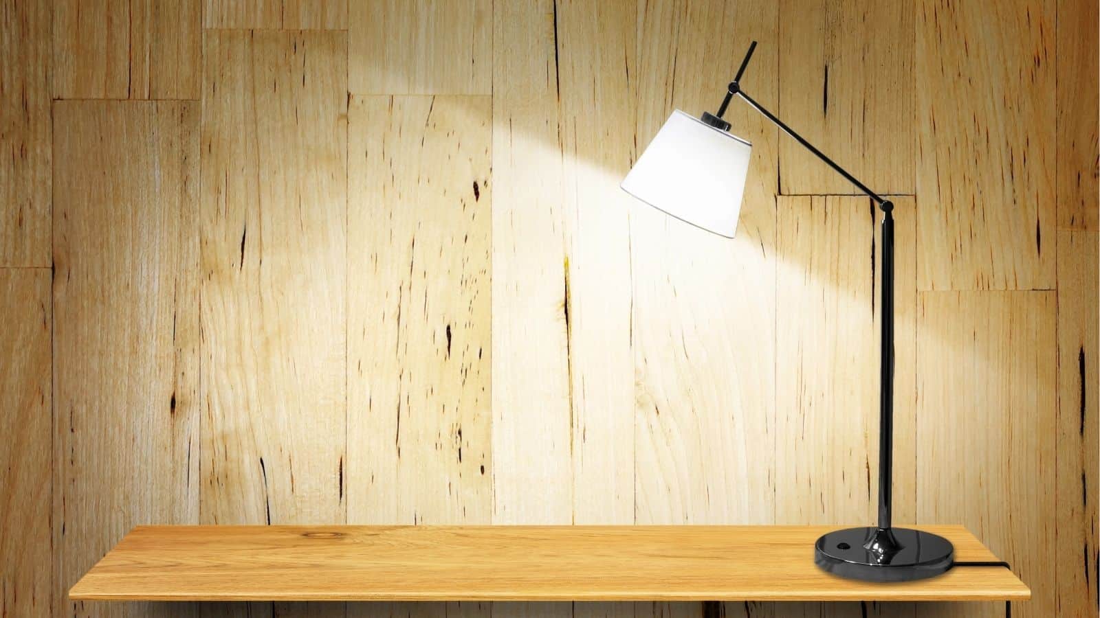 A modern black desk lamp with a white shade is turned on, casting light onto a wooden shelf in front of a light wood-paneled wall.