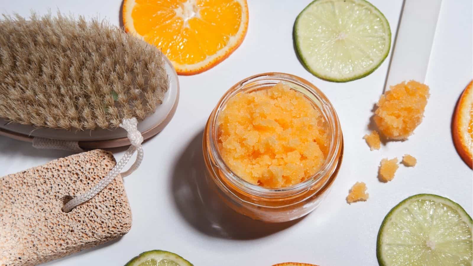 A jar of orange sugar scrub sits on a white surface surrounded by citrus slices, a dry brush, and a pumice stone.