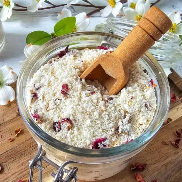 A glass jar of bath salts with dried flower petals sits on a wooden board, accompanied by a small wooden scoop and surrounded by white flowers.