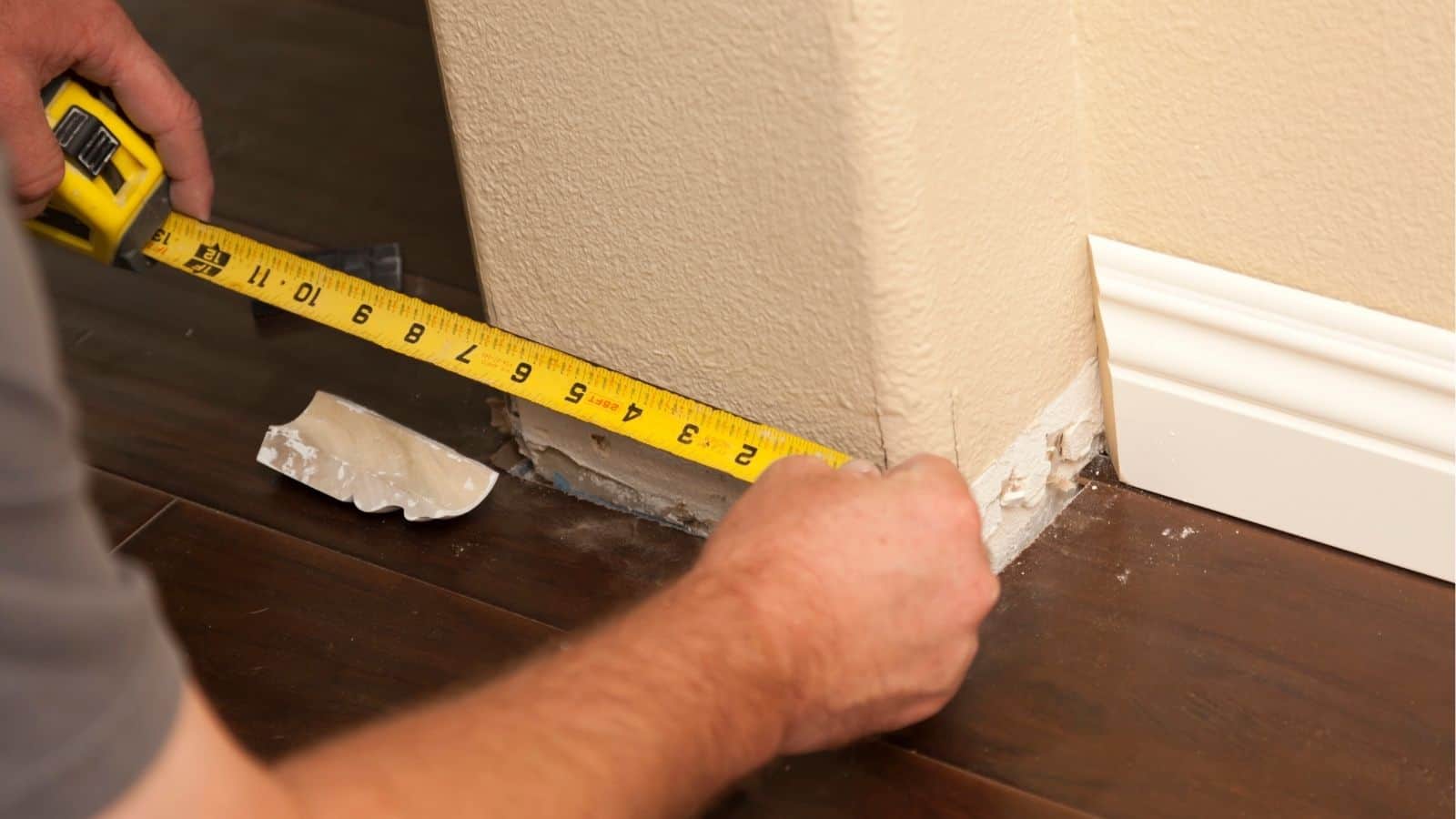 A person measures the length of baseboard space along a wall using a yellow tape measure on a dark wood floor.