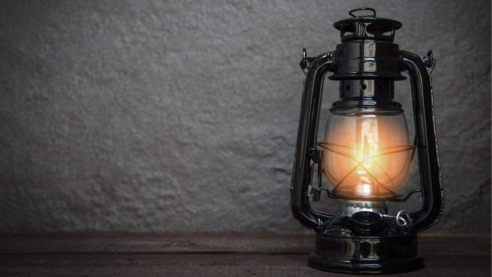 A lit vintage kerosene lantern sits on a wooden surface against a plain gray textured wall.