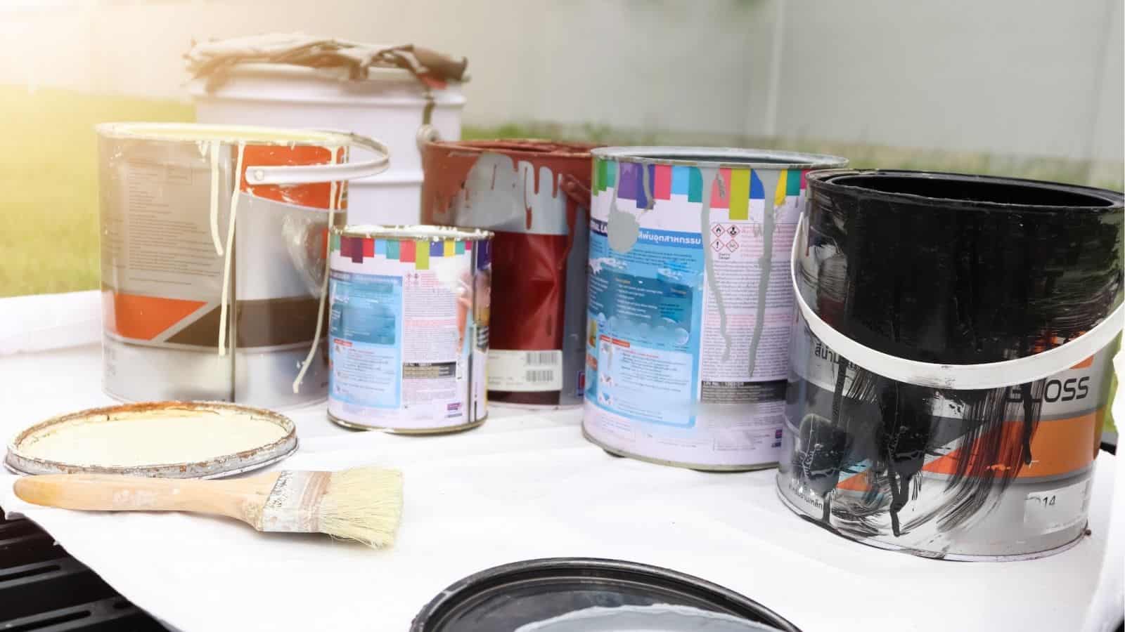 Several open and closed paint cans and a paintbrush are placed on a white surface outdoors, with splatters of paint visible on some cans.