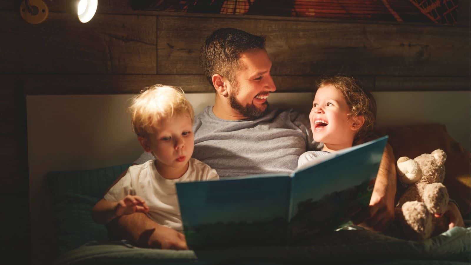 A man sitting in bed reads a storybook to two young children, one listening and smiling, the other looking at the book, with a stuffed animal beside them.