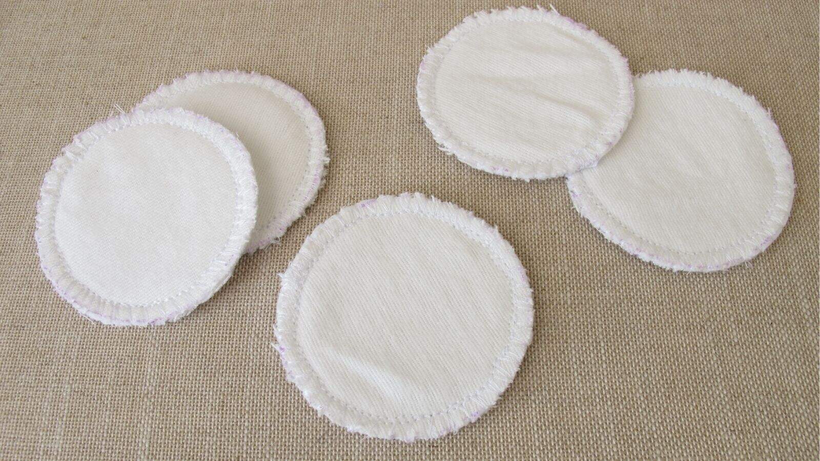 Five round, white reusable cotton pads with frayed edges are arranged on a beige fabric surface.
