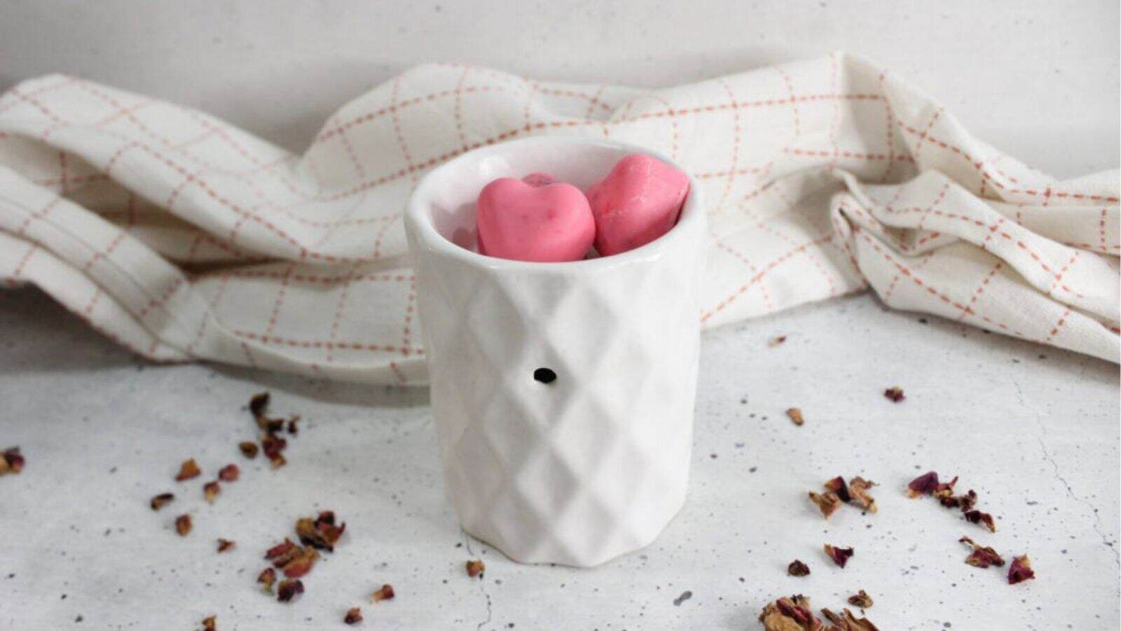 A white ceramic wax melt burner with two pink heart-shaped wax melts inside, set on a light surface with dried flower petals and a checkered cloth in the background.