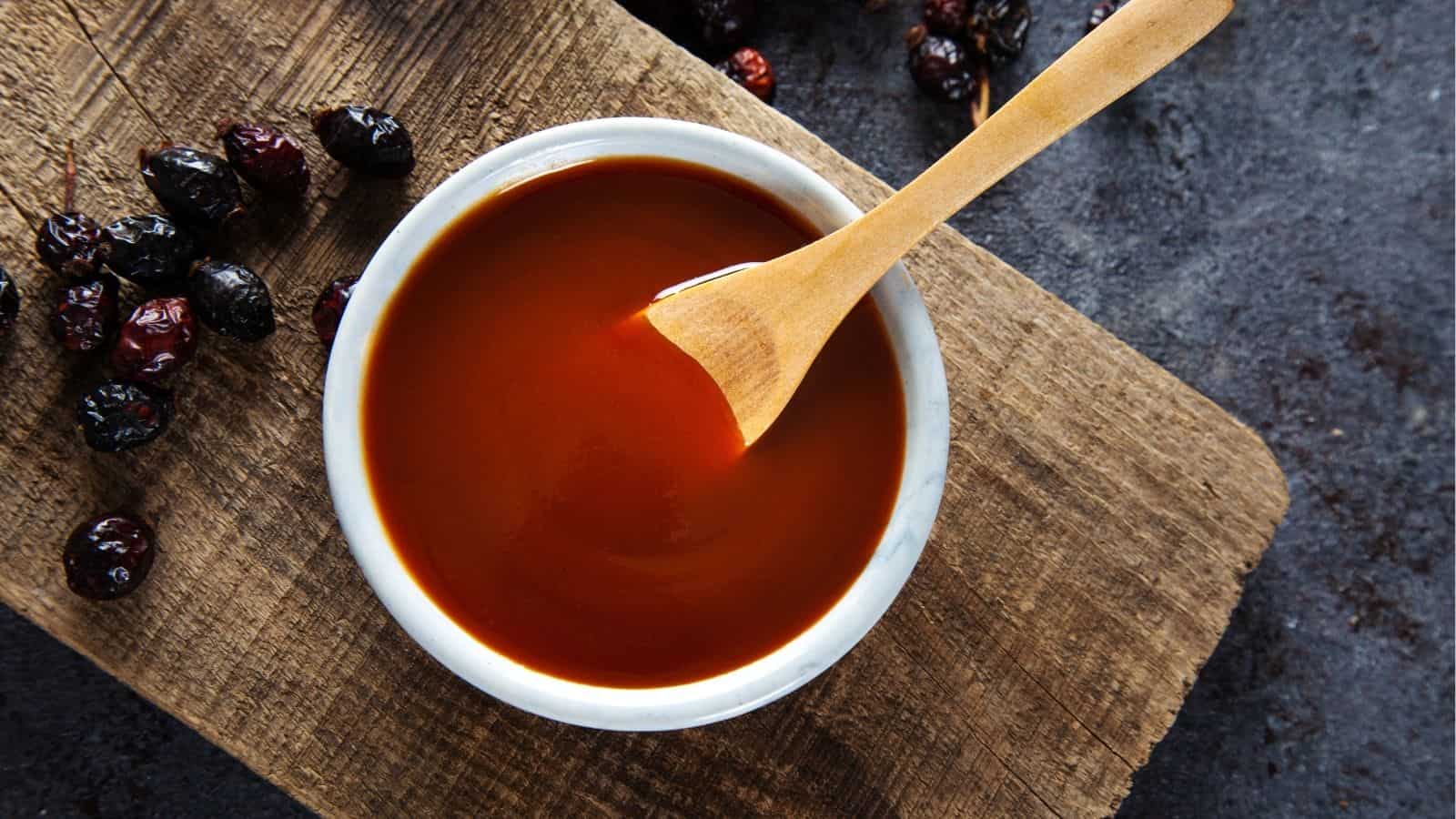 A small white bowl filled with reddish-brown sauce and a wooden spoon, placed on a wooden board with some dried berries nearby.