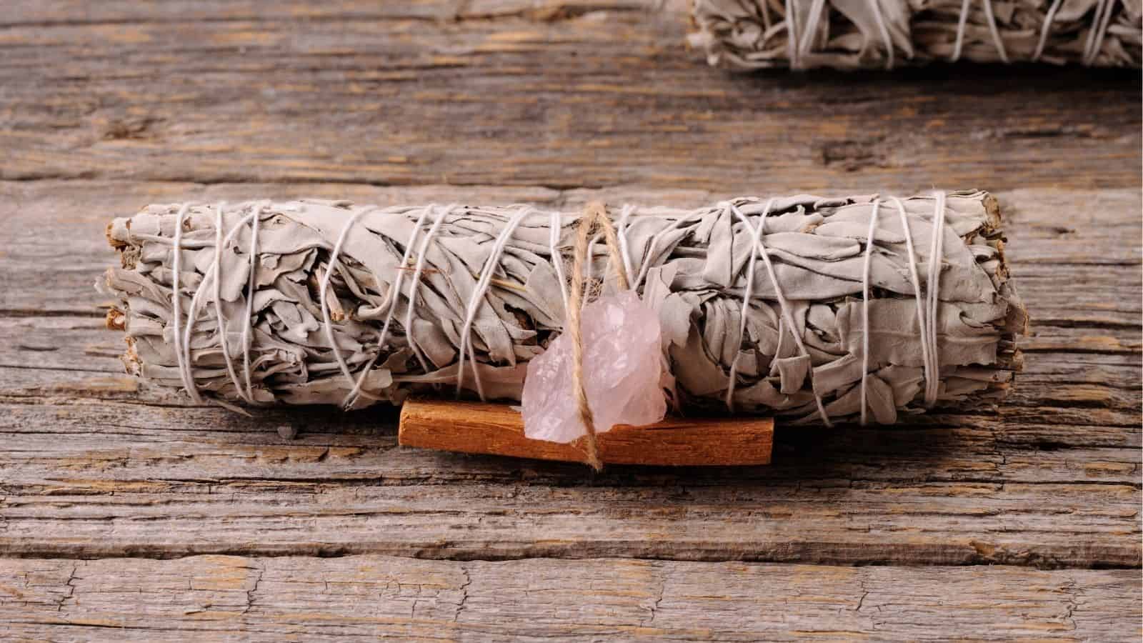 A bundle of dried sage wrapped with string, two rose quartz crystals, and a small stick of wood sit on a rustic wooden surface.
