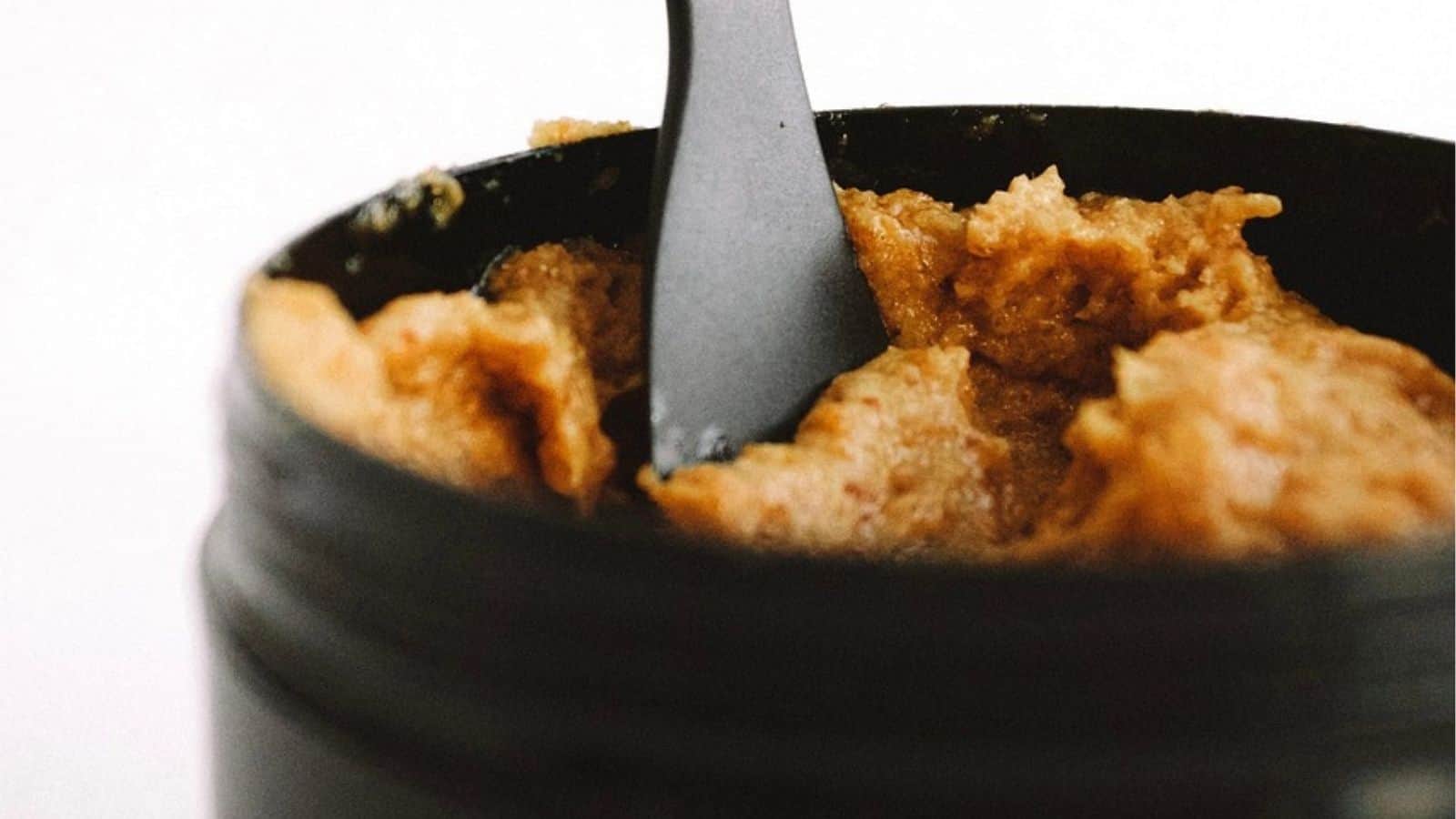 An open container filled with a thick, brown, paste-like substance being scooped by a black spoon.