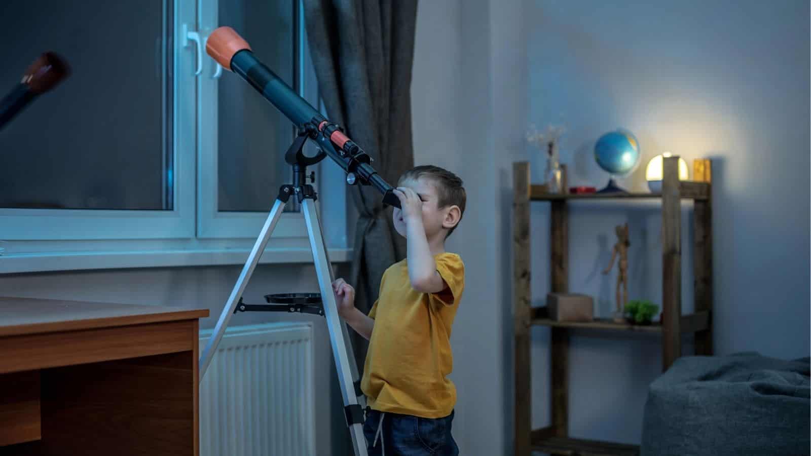 A young boy in a yellow shirt looks through a telescope by a window in a dimly lit room with shelves and a globe visible in the background.