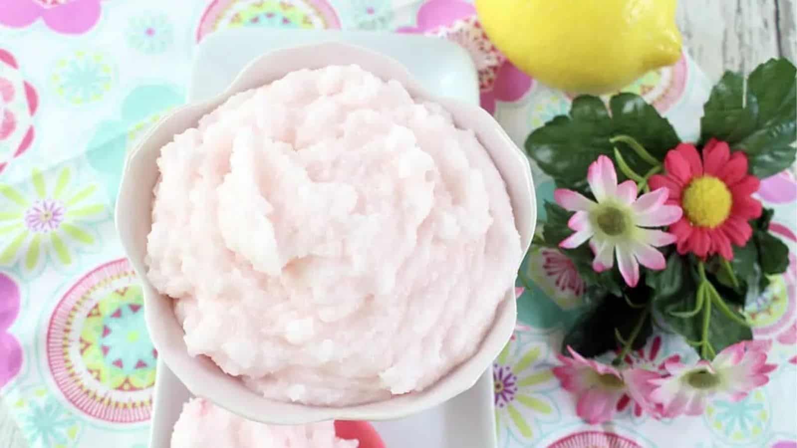 A bowl of light pink fluffy mixture sits on a floral-patterned surface next to a lemon and artificial flowers.