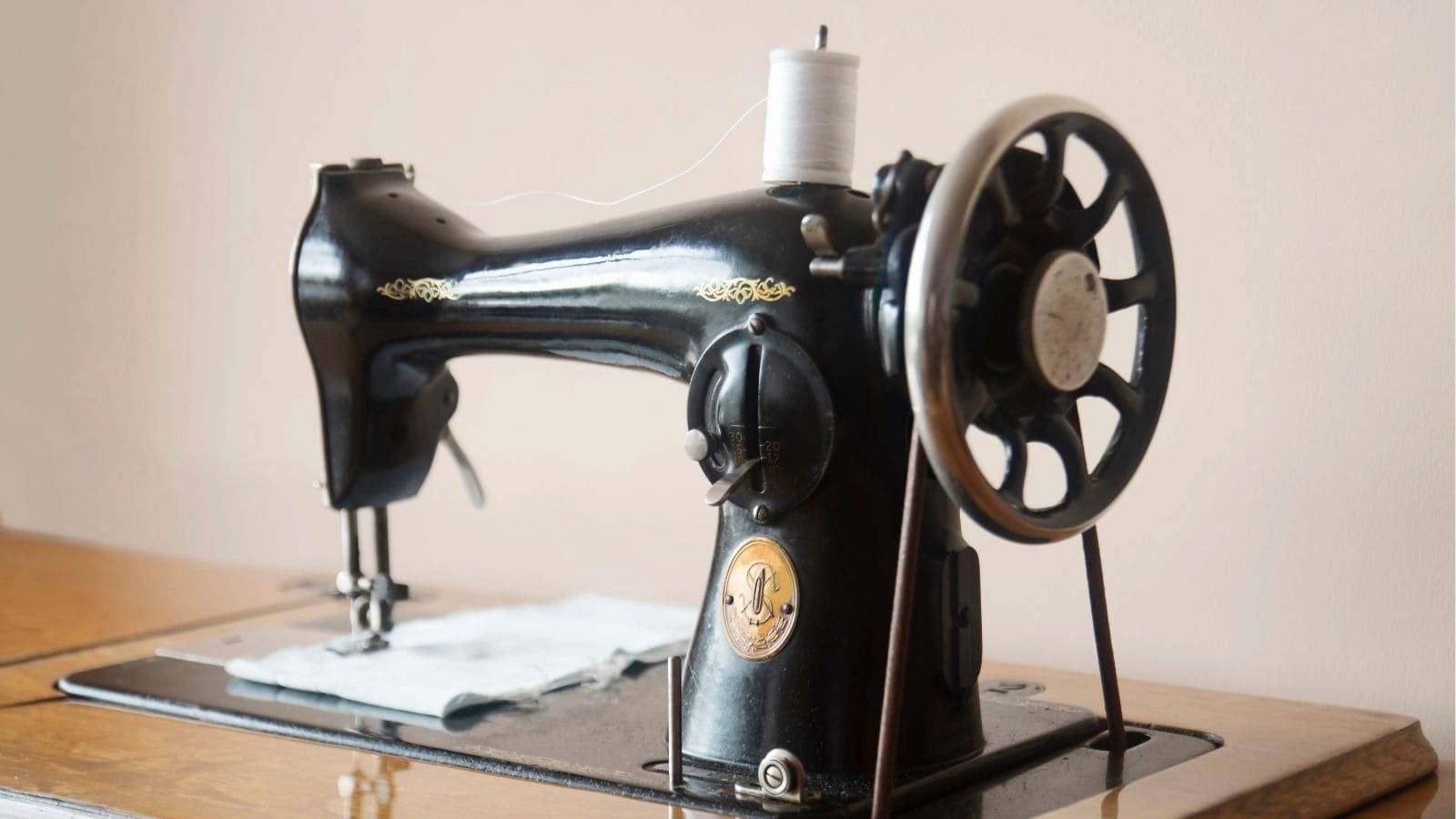 A vintage black sewing machine with a hand crank sits on a wooden table, threaded with white thread and holding a piece of fabric.