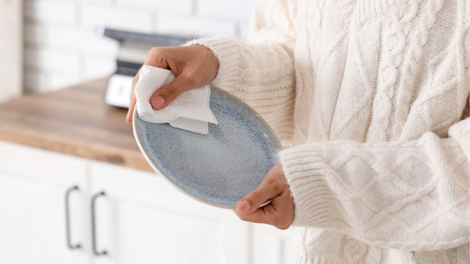 Person in a cream sweater drying a blue ceramic plate with a white cloth in a kitchen.