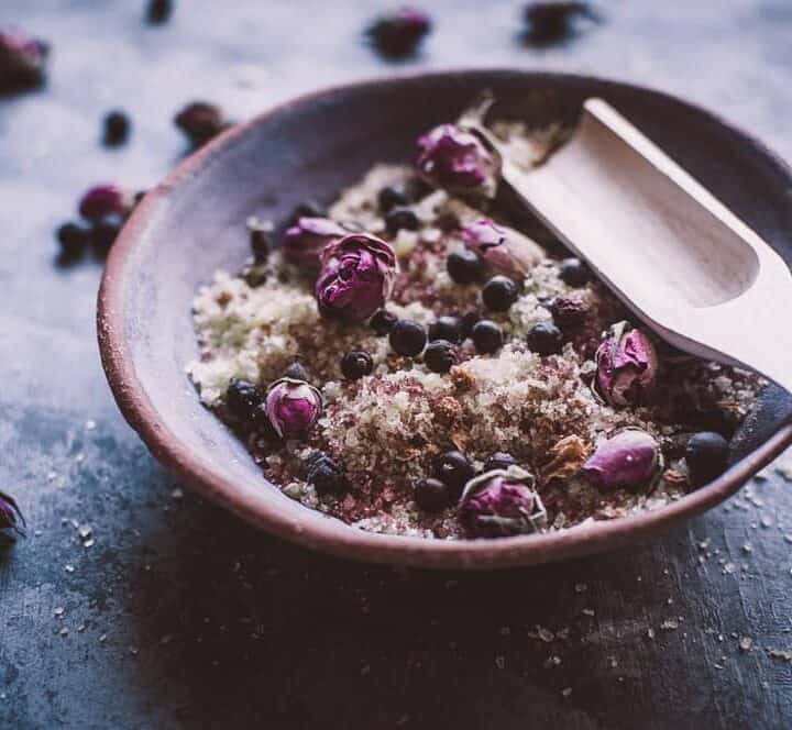 A ceramic bowl filled with a mixture of coarse salt, dried rosebuds, and berries, with a wooden scoop resting on the edge.