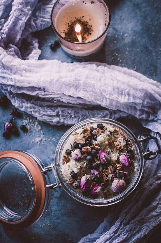 A glass jar filled with a mixture of dried flowers and herbs sits on a blue surface next to an open lid, with a lit candle and gauzy fabric in the background.