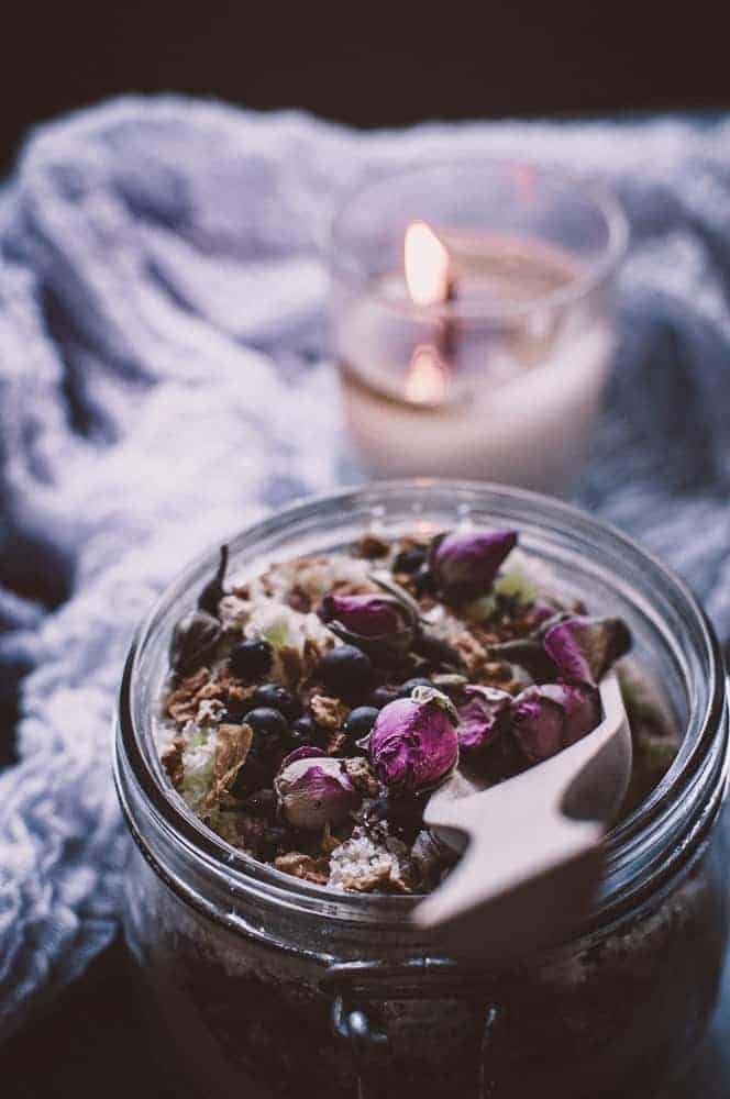 A glass jar filled with a mixture of dried flowers, herbs, and oats, with a wooden scoop on top; a lit candle and a textured cloth are in the background.